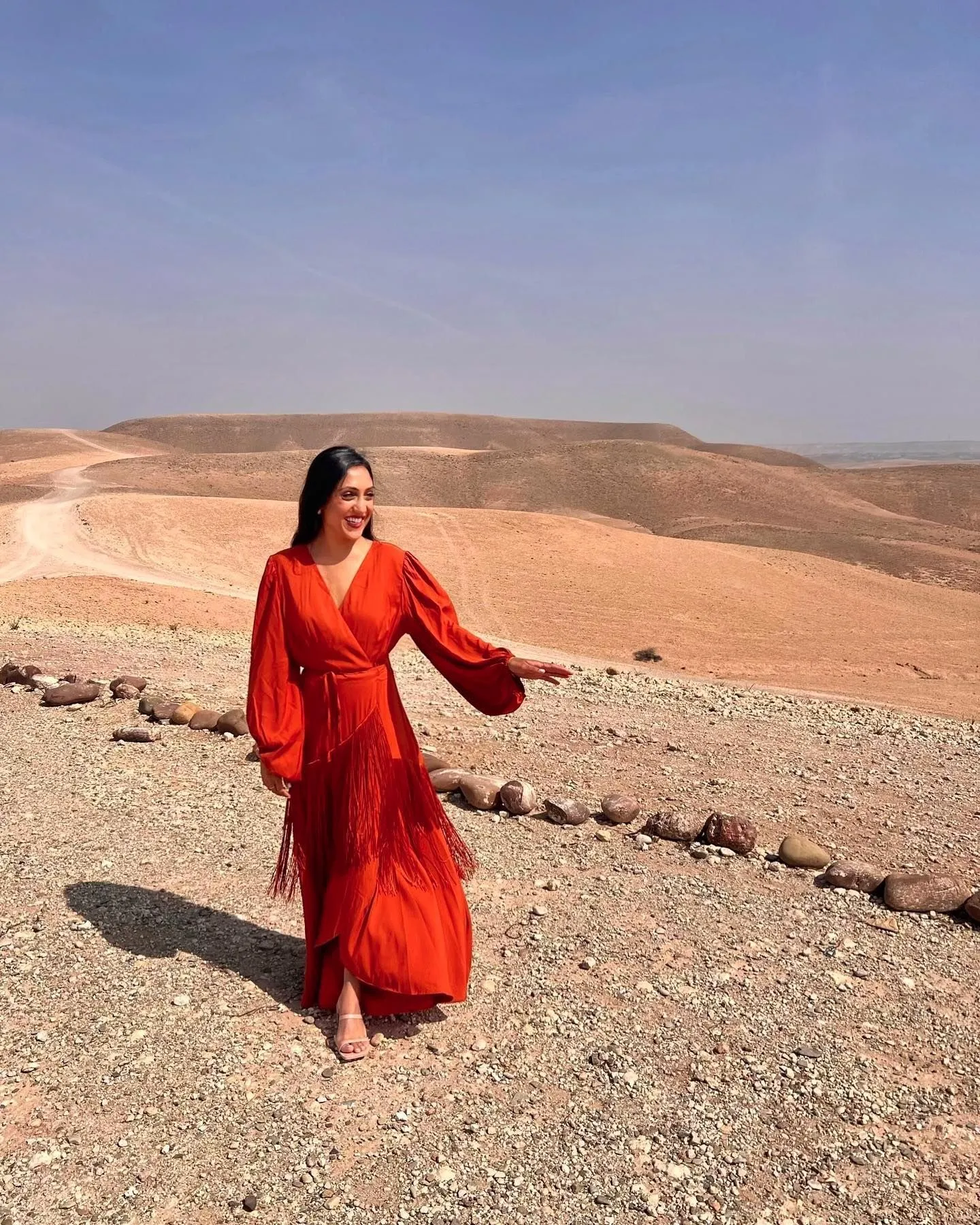 Model wearing a flowy rust-colored gauze maxi dress on a sand dune during sunset
