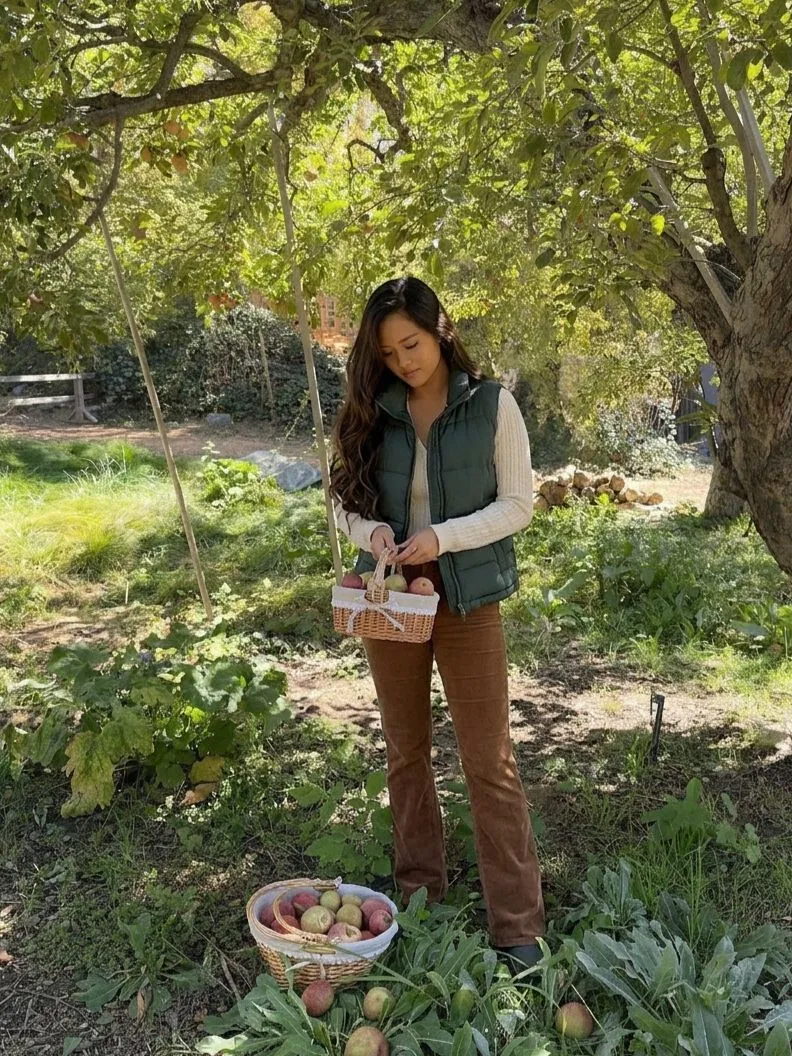Woman wearing a green puffer vest and brown bootcut corduroy pants in an orchard